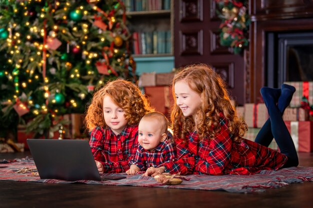 Crianças ruivas engraçadas ao lado da árvore de Natal e dos presentes se comunicam online por meio de um laptop.