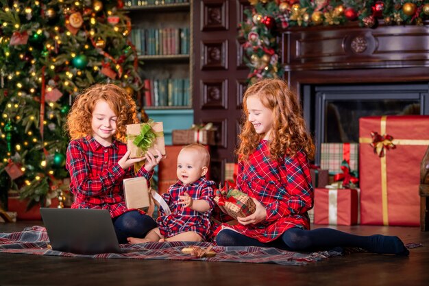 Crianças ruivas engraçadas ao lado da árvore de Natal e dos presentes se comunicam online por meio de um laptop.