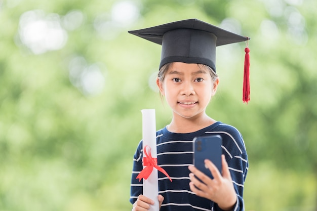 Crianças se formam com chapéu de formatura e certificado de graduação enrolado segurando um smartphone para ligar