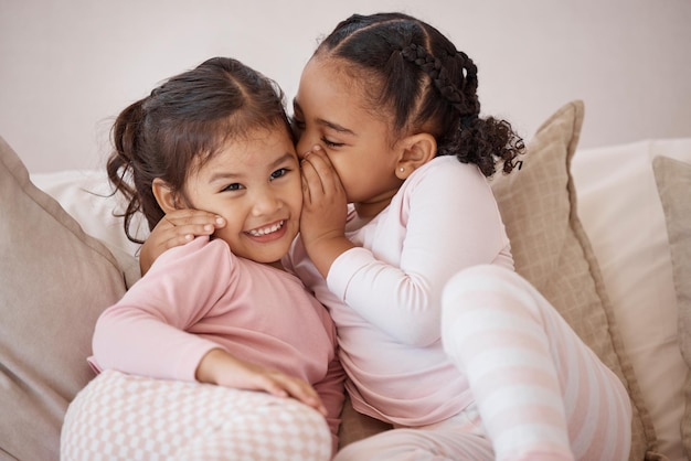 Crianças secretas e meninas sussurrando no ouvido com amor, confiança e felicidade enquanto brincavam em um sofá em casa Adoção inter-racial e vínculo entre irmãs irmãs amigas e crianças se divertindo