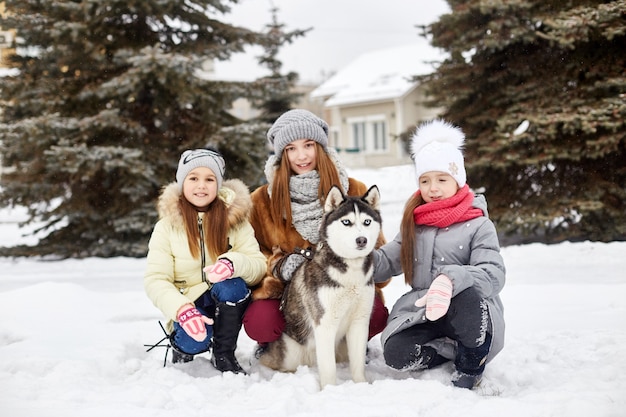 Crianças sentam na neve e acariciam o cão husky