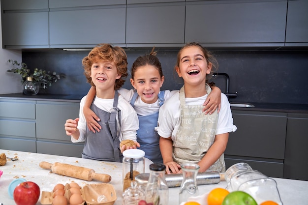 Crianças sorridentes de avental olhando para a câmera na mesa com massa de biscoito e vários ingredientes na cozinha leve em casa