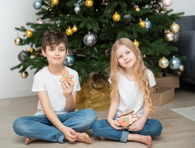 Crianças - um menino e uma menina comendo pão de mel de Natal perto da árvore de Natal na sala de estar