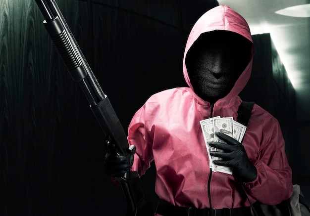 Criminoso de uniforme rosa e máscara escondida segurando a espingarda e carregando um saco de dinheiro após o assalto à casa