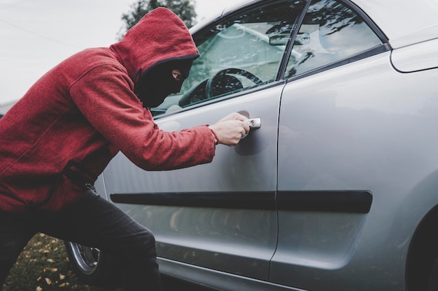 Foto criminoso em balaclava preta e capuz abre o veículo de alguém com a chave mestra carro quebrando por homem desconhecido com rosto escondido jovem abre a fechadura do carro para entrar forçando a porta do carro