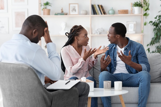 Crise do casamento. Jovens cônjuges negros discutindo na sessão de terapia familiar, sentados no sofá no escritório do conselheiro, espaço livre