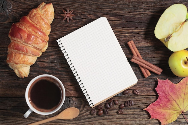 Croissant con café, libreta abierta y manzana en la mesa de madera oscura, vista superior