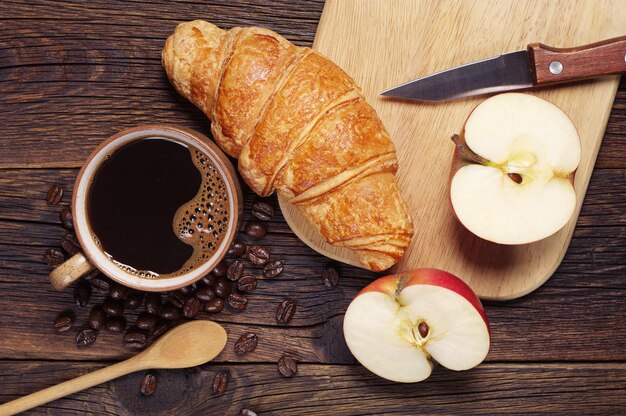 Croissant, café negro y manzana para desayunar en una mesa de madera oscura.