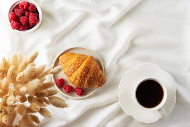 Croissant crujiente francés con frambuesas y una taza de café en la cama