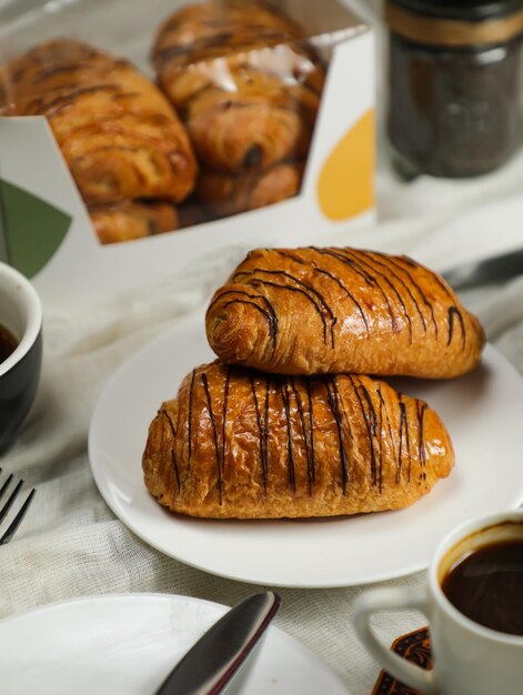 Foto croissant de chocolate servido em prato com uma xícara de café preto com faca e garfo isolados no guardanapo vista lateral do café da manhã francês alimento assado em fundo cinza