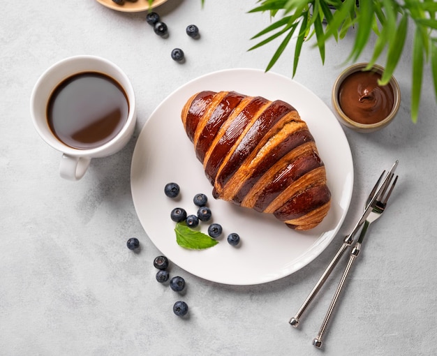 Croissant fresco con chocolate en un plato blanco sobre un fondo claro con una taza de café de arándanos y hoja de palma Delicioso concepto de desayuno casero