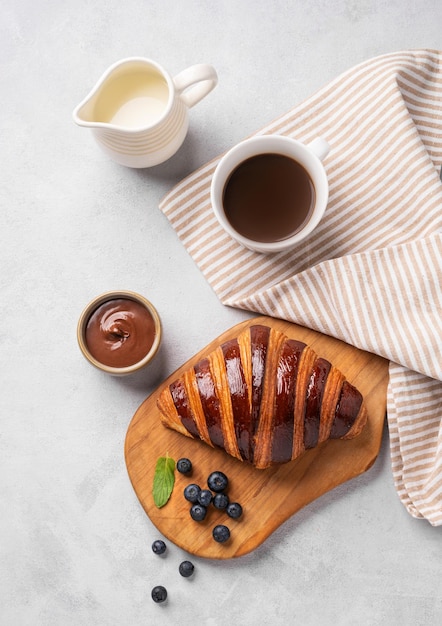 Croissant fresco con chocolate en una tabla de corte en un fondo claro con una taza de café de arándanos y una jarra de leche Delicioso concepto de desayuno casero