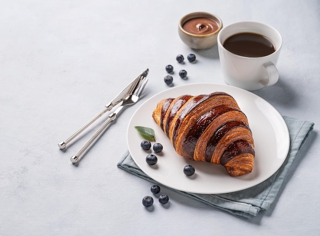 Croissant fresco com chocolate e mirtilos em um prato branco sobre um fundo claro com uma xícara de café preto Delicioso café da manhã caseiro conceito