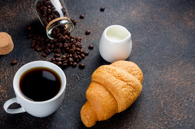 Croissant fresco com uma caneca de café preto e grãos de café no fundo escuro de concreto.