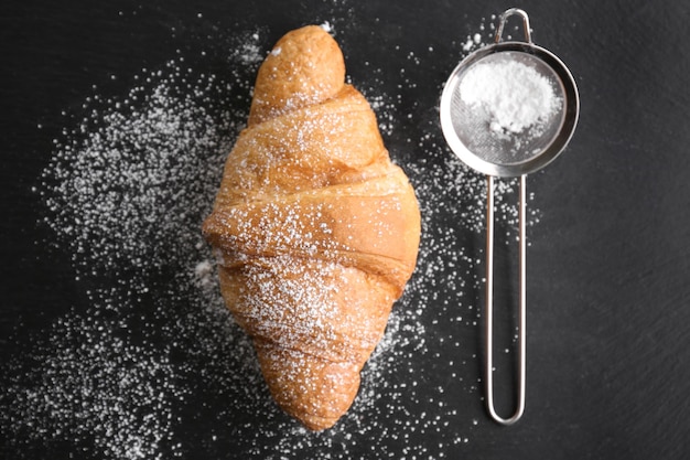 Croissant fresco y delicioso y tamiz con azúcar en polvo sobre fondo oscuro