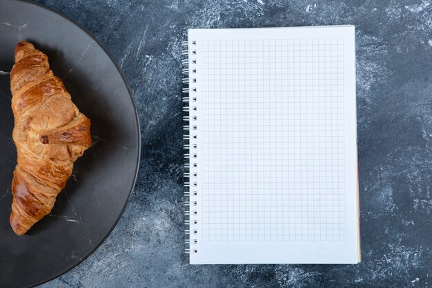 Croissant fresco y sabroso con cuaderno abierto colocado sobre la mesa de piedra.