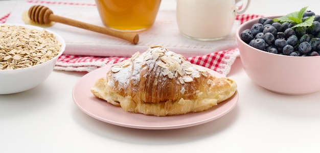 Croissant horneado espolvoreado con azúcar en polvo, arándanos y avena en un plato de cerámica sobre una mesa blanca, desayuno