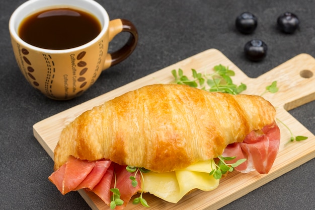 Croissant con jamón y queso y verduras en la tabla de cortar. Taza de cafe. Vista superior. Superficie negra.