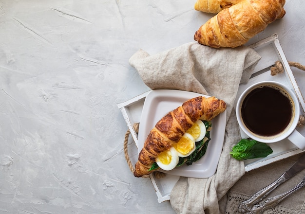 Croissant mit Ei und sautiertem Spinat und Kaffee in Holztablett auf hellgrauem Beton.