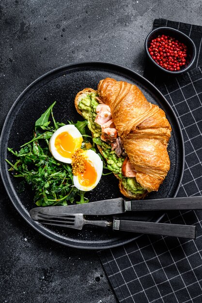 Croissant mit heiß geräuchertem Lachs, Avacado, Rucola und Ei. Schwarzer Hintergrund. Draufsicht