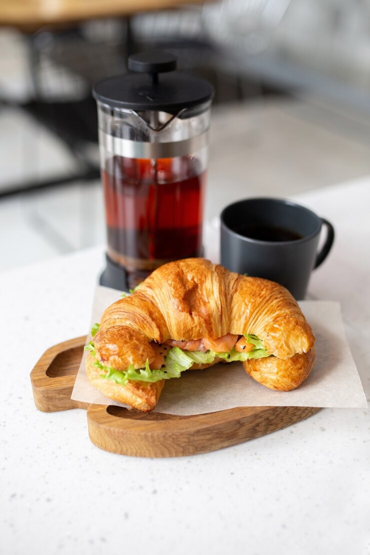 Croissant mit Lachs und frischem Salat wird zum Mittagessen auf einem Holzbrett serviert. Schwarzer Tee in einer Teekanne. Fitness Essen in einem Café.
