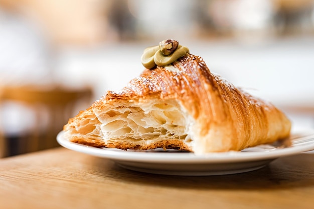 Un croissant de pistacho mordido con relleno de crema en un plato blanco en la mesa de madera de una panadería Fondo Bokeh Foto de estilo de vida enfoque selectivo profundidad de campo