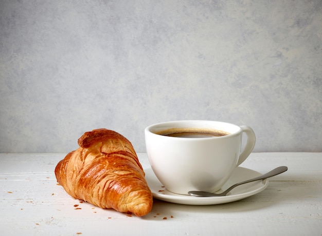 Croissant recién horneado y taza de café en la mesa de madera blanca