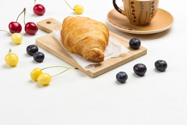 Croissant, Tasse Kaffee, Kirschen und Blaubeeren auf dem Tisch.