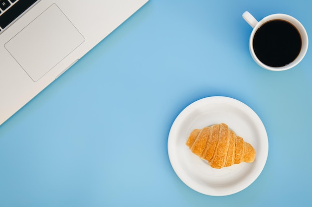 Croissant Tasse Kaffee und Laptop auf blauem Hintergrund flach gelegt