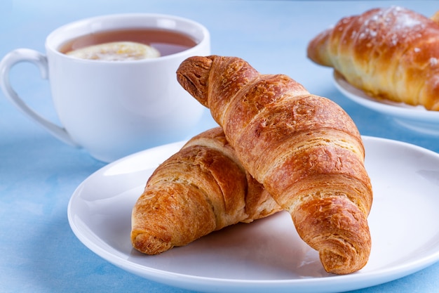 Croissants acabados de fazer e uma xícara de chá quente com limão no café da manhã francês em um fundo azul