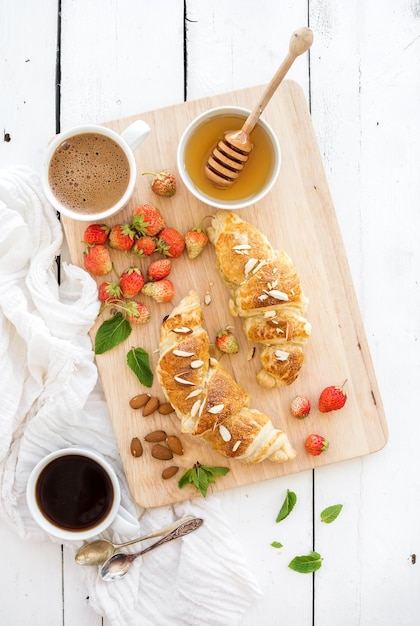 Croissants de almendras recién horneados con fresas de jardín, miel de menta y café en una tabla para servir sobre fondo blanco de madera rústica