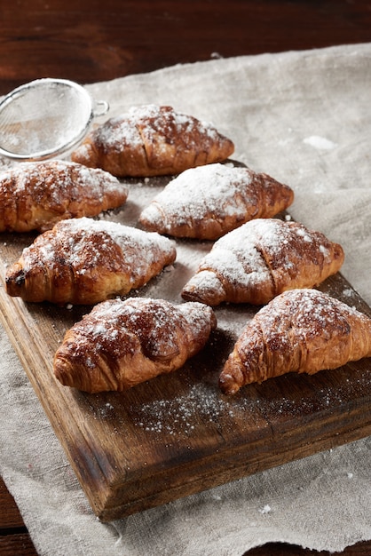 Croissants assados polvilhados com açúcar de confeiteiro repousam sobre uma placa de madeira marrom