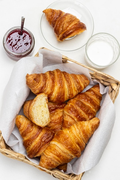 Croissants en cesta de mimbre. Vaso de leche y tarro de mermelada. Endecha plana. Fondo blanco.