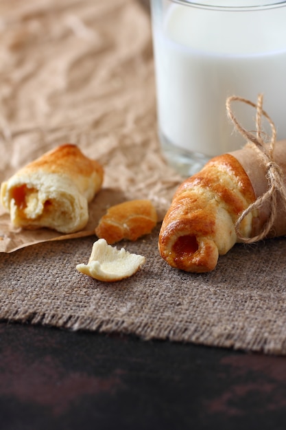 Croissants com geléia amarrados com corda quebrada e um copo de leite em papel kraft e toalha de mesa de estopa.