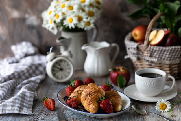 Croissants con fresas rojas frescas con una taza de café en una mesa de madera y ramo de margaritas en una jarra.