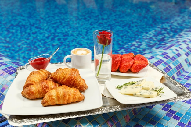 Croissants, Marmelade, Käse, Erdbeere und Kaffee stehen auf einem Tablett am Rand eines Swimmingpools. Konzept der Hotellerie