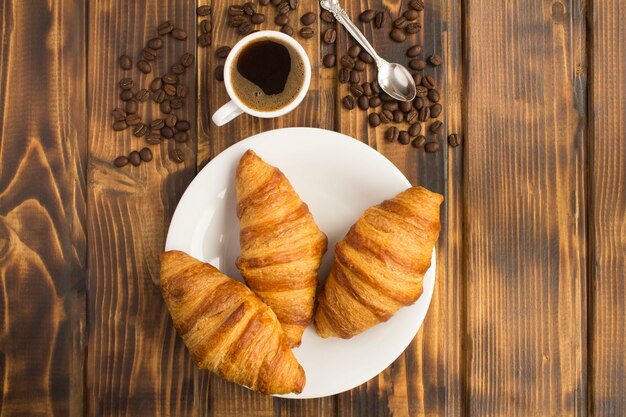 Croissants en el plato blanco y café negro en la taza sobre el fondo de madera marrón. Vista superior. Copie el espacio.