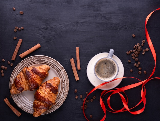 Croissants en un plato de cerámica, una taza de café, cinta roja, palitos de canela y granos de café sobre una mesa de madera negra.