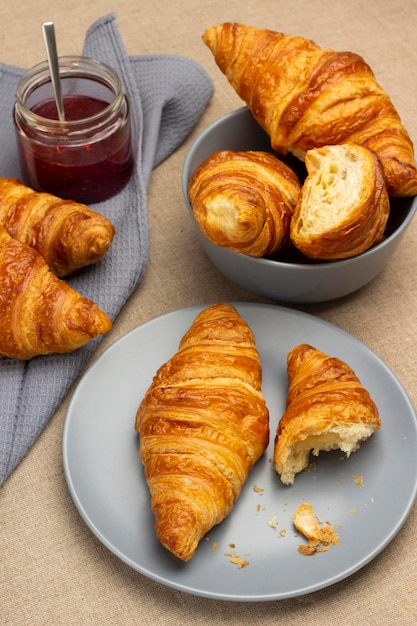 Croissants en plato gris y tazón. Tarro con mermelada en servilleta gris. Ropa de arpillera en la mesa. Vista superior.