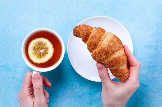 Croissants recién horneados y una taza de té caliente con limón en la mano para el desayuno francés sobre un fondo azul.