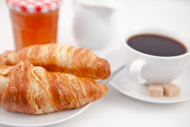 Croissants y una taza de café en platos blancos con leche de azúcar y una olla de mermelada