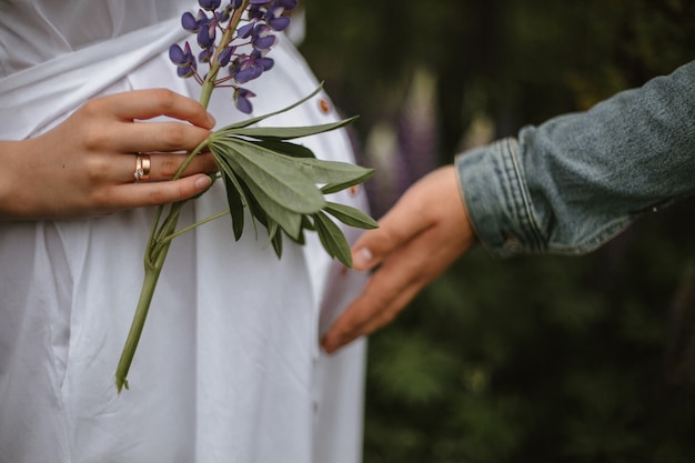 Crop-Ansicht einer schwangeren Frau und einer zarten Hand, die violette Lupine mit Ehering hält und ein Mann, der den Bauch mit dem Kind berührt