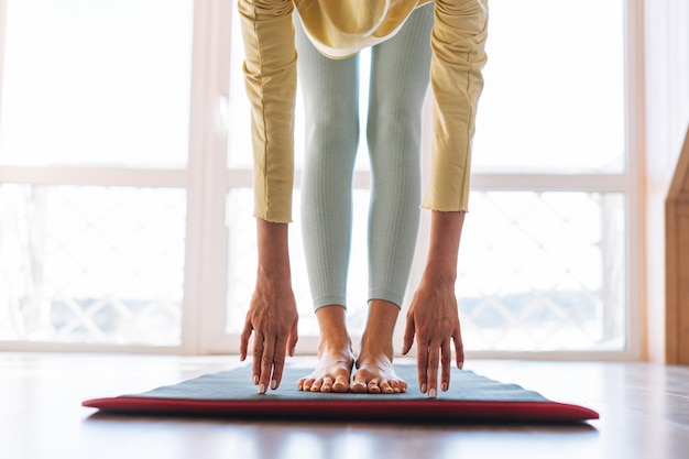 Crop-Foto-Beine einer jungen, schlanken, blonden Fitness-Frau auf einer Sportmatte, die Morgenübungen in der Nähe des Fensters des Hauses macht