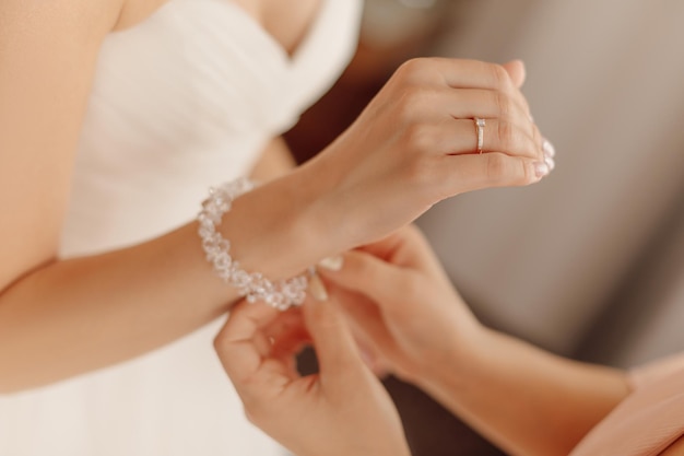 Crop-Foto von Händen einer nicht erkennbaren Frau in Beige, möglicherweise Mutter, Freundin oder Schwester, die die Braut in Weiß für den Hochzeitstag vorbereitet und dabei hilft, das Armband an ihrer Hand zu befestigen. Morgenversammlung der Braut