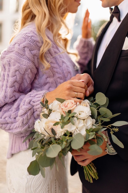 Crop-Foto von nicht erkennbaren Personen, möglicherweise blonde Braut, die eine lila Jacke und ein weißes Kleid und einen Ehering trägt, und ein Bräutigam in einem schwarzen Anzug, der nah beieinander steht und einen Strauß weißer und rosa Rosen hält