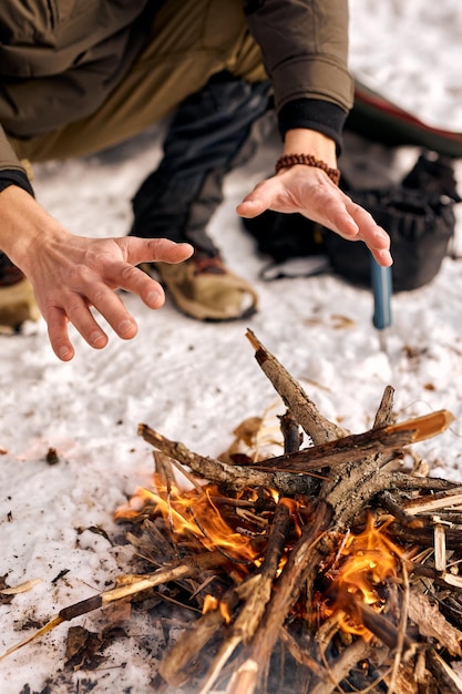 Cropped Mann warme Hände durch Feuer sitzen neben Feuer Reisekonzept Cropped Mann genießen Sie die Erkundung neuer Orte camping Brennen von Holz am Abend in der Natur im Winterwald allein Nahaufnahme Hände