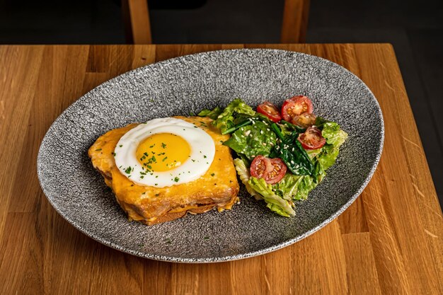 Croque madame café da manhã servido em um prato cinza com mistura de salada e tomate cereja para guarnição em uma mesa de madeira em um restaurante