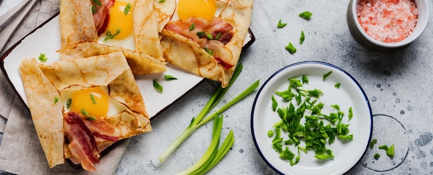 Crpe mit Spiegelei, Käse, Speck und Frühlingszwiebeln zum Frühstück auf hellgrauem Betonhintergrund.