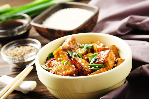 Crujientes cubos de tofu salteados con cebollino en un plato de arcilla sobre una mesa de cocina de madera con una servilleta de cebollino, pimienta y salsa de soja a un lado