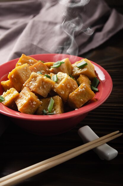 Crujientes cubos de tofu salteados con cebollino en un plato de arcilla sobre una mesa de cocina de madera con una servilleta a un lado